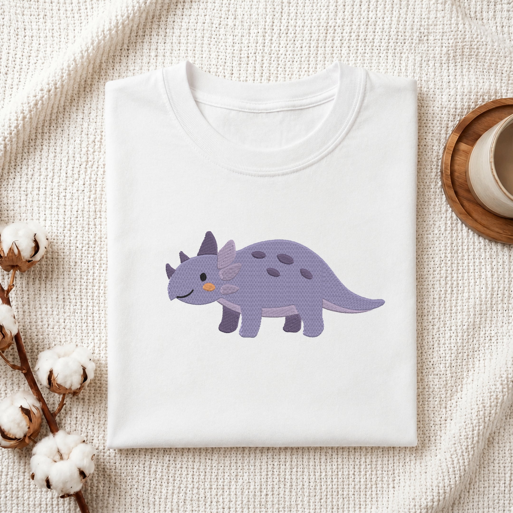 A folded white crewneck t-shirt featuring a cute cartoon Triceratops dinosaur graphic in purple, displayed on a textured cream knit blanket with decorative cotton bolls and a wooden cup.