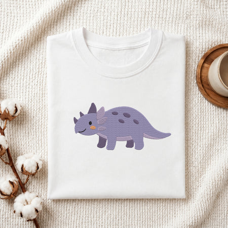 A folded white crewneck t-shirt featuring a cute cartoon Triceratops dinosaur graphic in purple, displayed on a textured cream knit blanket with decorative cotton bolls and a wooden cup.