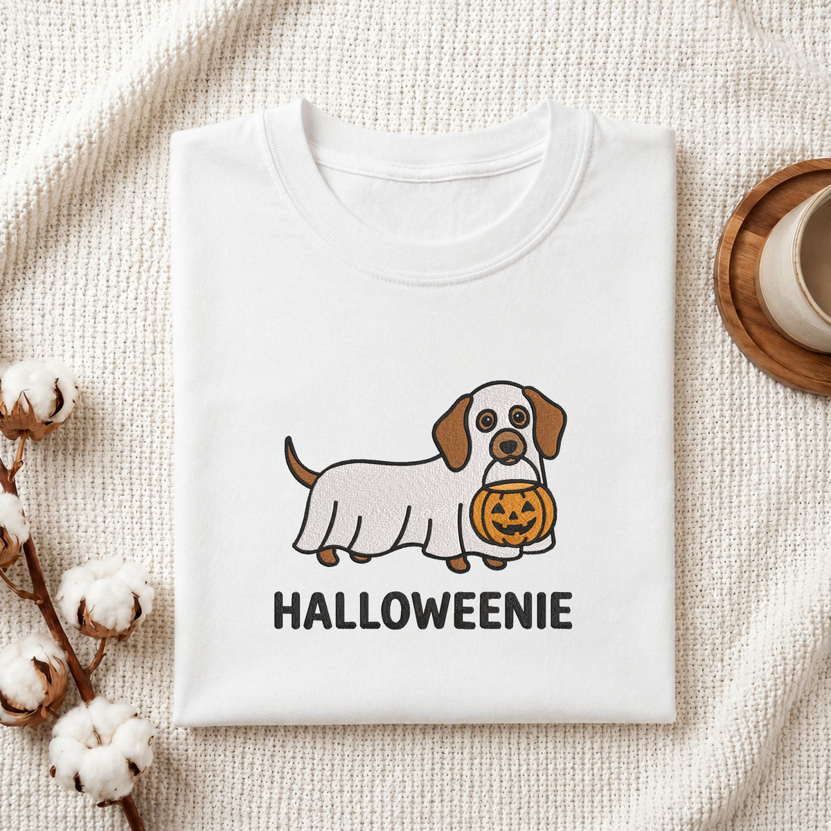 A folded white T-shirt with a cute cartoon dog wearing a Halloween cover and holding a jack-o'-lantern, with the text 'HALLOWEENIE' printed below. The shirt is styled on a textured cream fabric with cotton bolls and a wooden cup in the background.