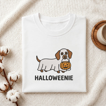 A folded white T-shirt with a cute cartoon dog wearing a Halloween cover and holding a jack-o'-lantern, with the text 'HALLOWEENIE' printed below. The shirt is styled on a textured cream fabric with cotton bolls and a wooden cup in the background.