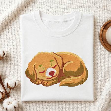 A folded white crewneck t-shirt with a graphic of a sleeping golden retriever dog, styled on a textured cream fabric with cotton bolls and a wooden tray.