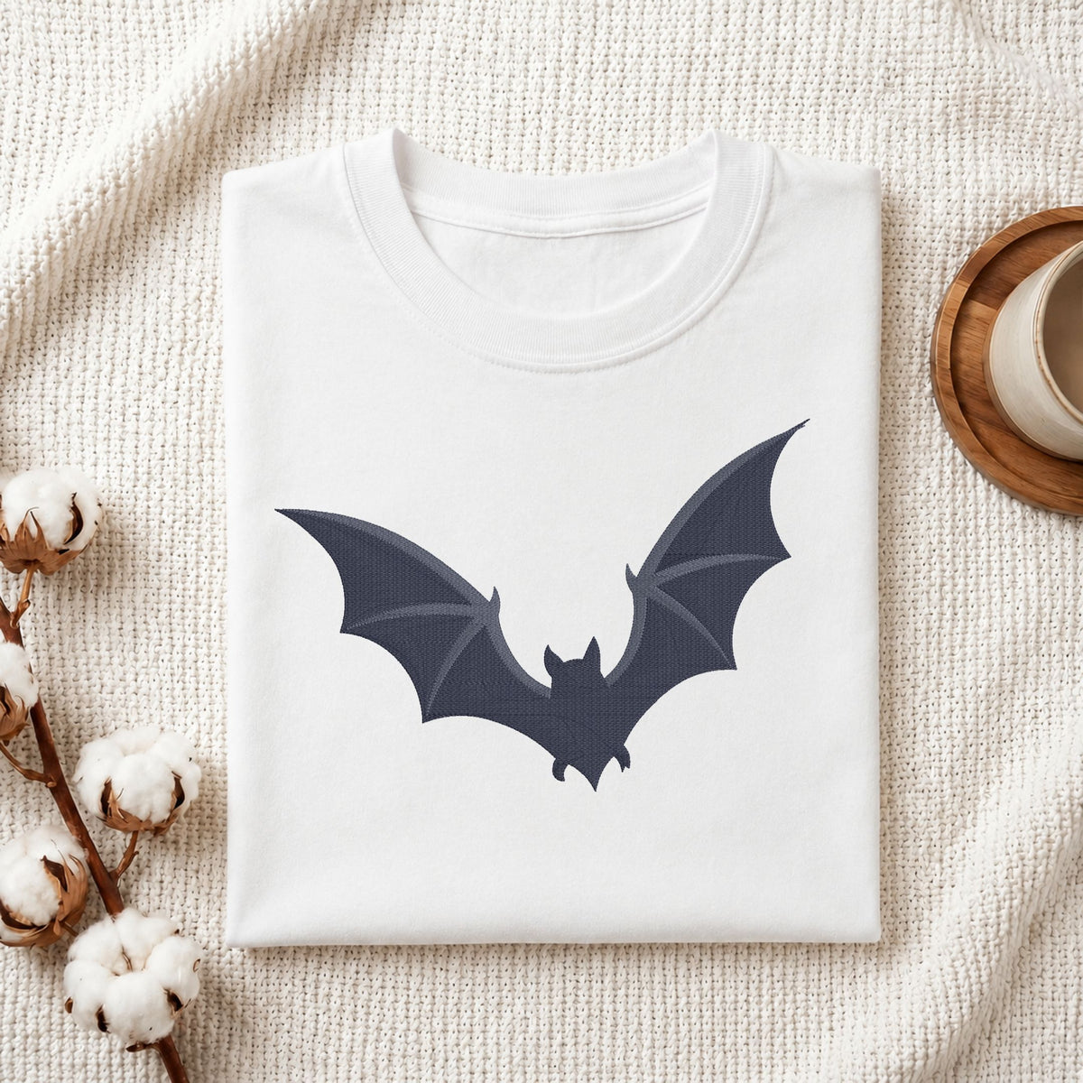 A folded white crewneck t-shirt featuring a dark navy bat graphic on the front, styled on a textured cream knit blanket with cotton bolls and a wooden cup in the background.