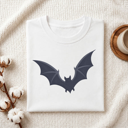 A folded white crewneck t-shirt featuring a dark navy bat graphic on the front, styled on a textured cream knit blanket with cotton bolls and a wooden cup in the background.