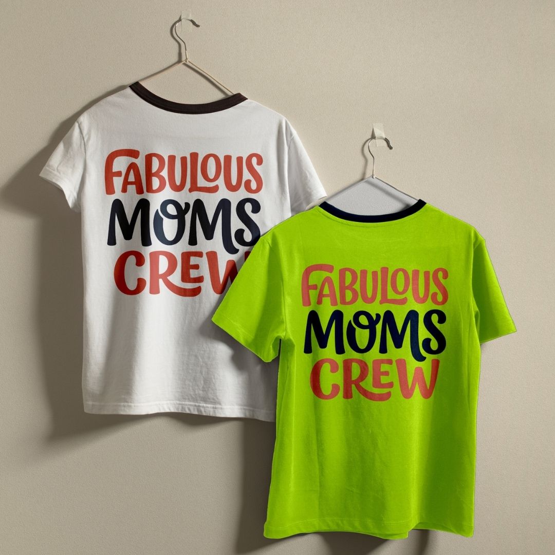 Two t-shirts hanging on clothing hangers against a neutral wall: one white t-shirt with black trim around the neck and one vibrant neon green t-shirt with navy trim, both featuring the "FABULOUS MOMS CREW" graphic design.