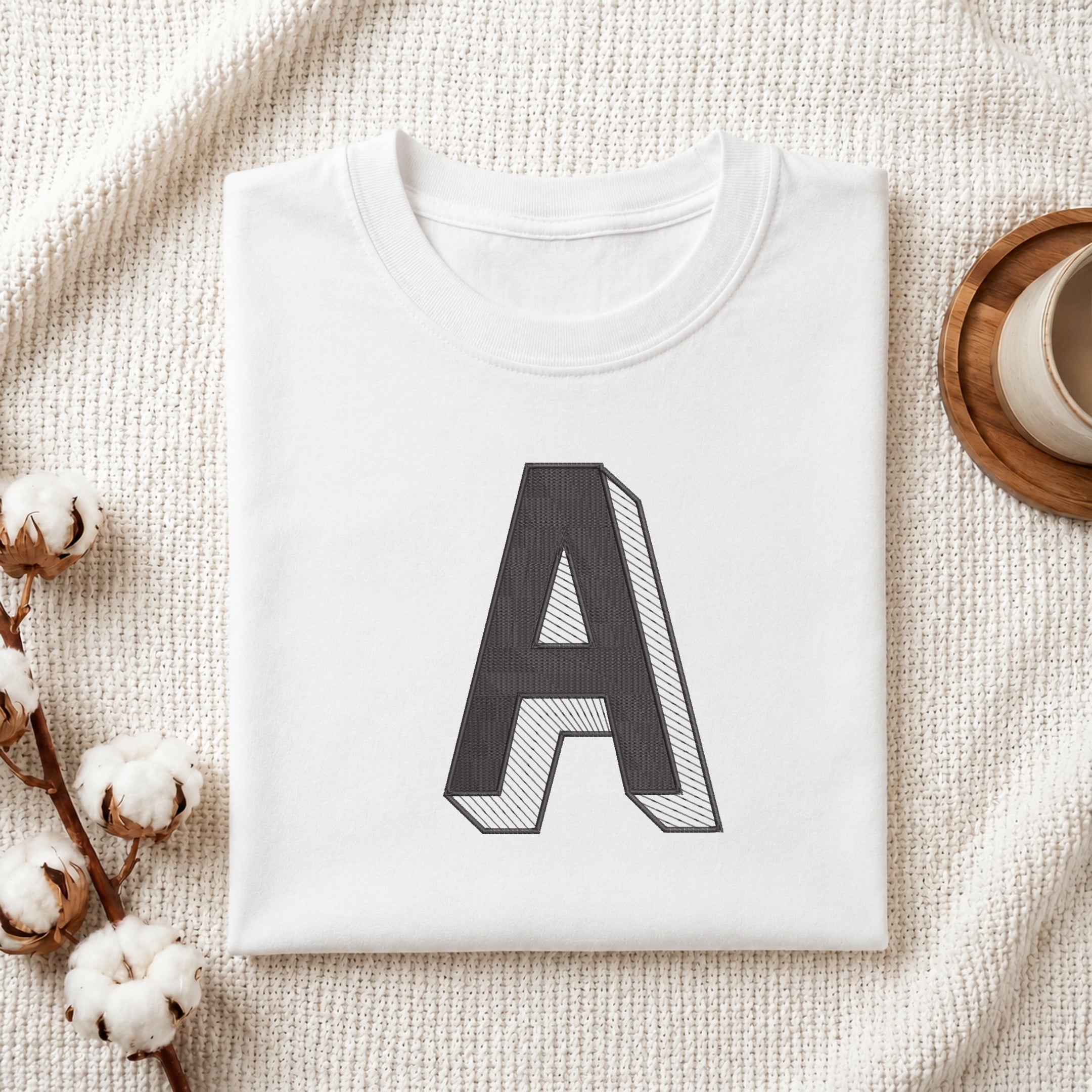 A folded white t-shirt with a large black block letter 'A' printed on the front, styled on a textured cream blanket with cotton bolls and a wooden cup in the background.