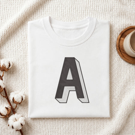 A folded white t-shirt with a large black block letter 'A' printed on the front, styled on a textured cream blanket with cotton bolls and a wooden cup in the background.