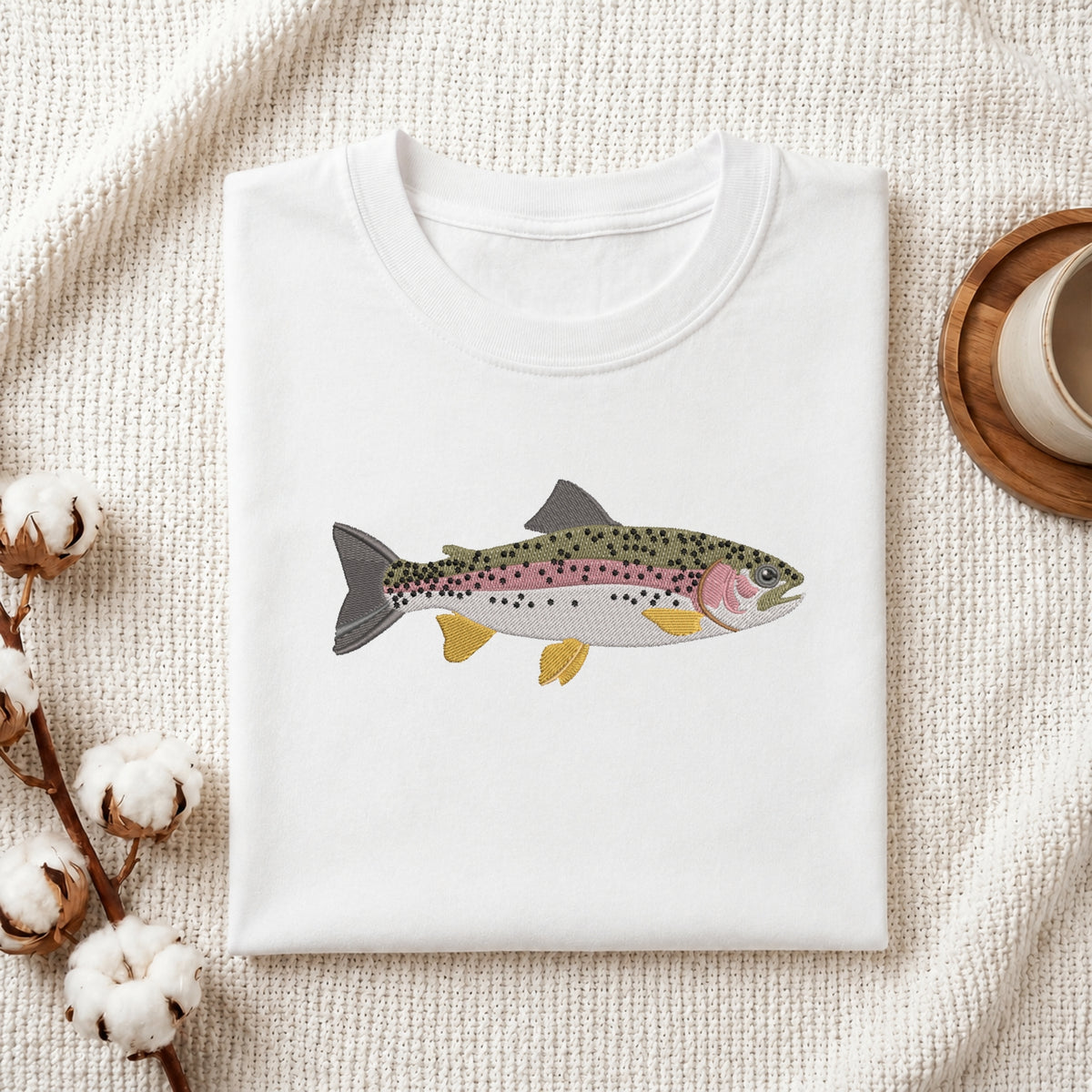 A folded white crewneck t-shirt featuring a detailed illustration of a rainbow trout on the chest, styled on a textured cream fabric with cotton bolls and a wooden cup.