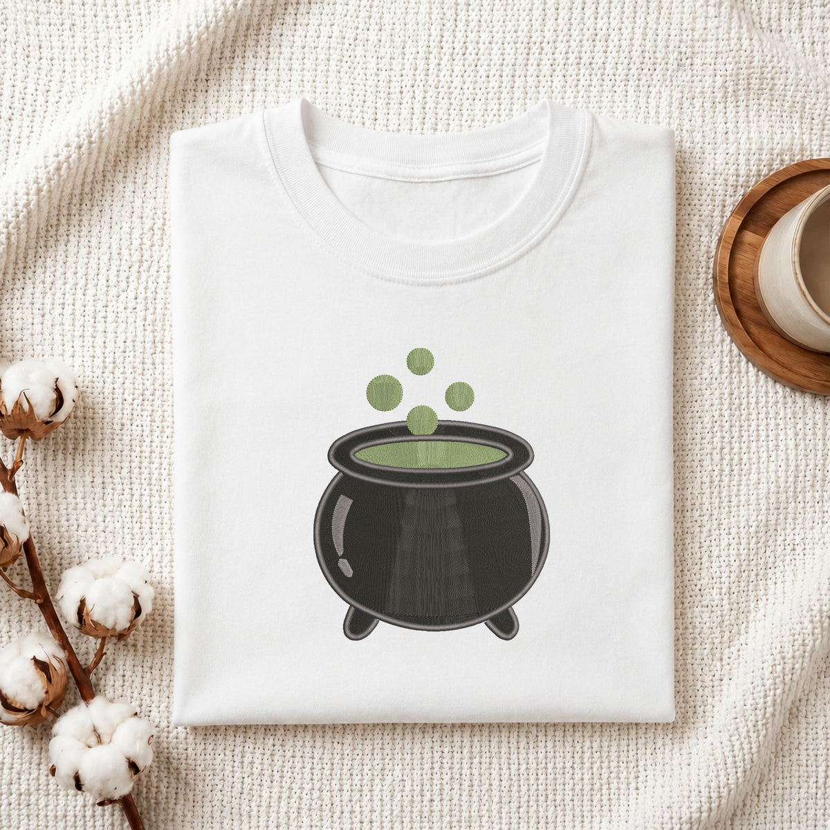 Folded white cotton t-shirt mockup featuring a witch cauldron machine embroidery design, styled with cotton stems and a wooden cup.
