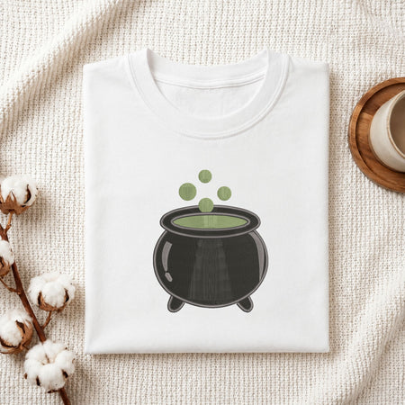 Folded white cotton t-shirt mockup featuring a witch cauldron machine embroidery design, styled with cotton stems and a wooden cup.