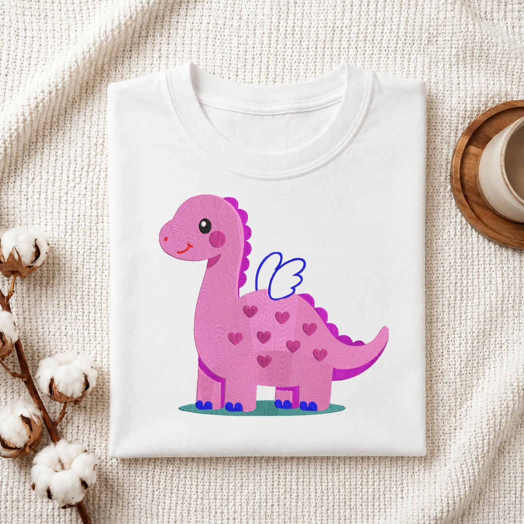 A folded white crewneck t-shirt featuring a cute pink cartoon dinosaur with blue wings and pink hearts, styled on a textured beige fabric with cotton bolls and a wooden cup.