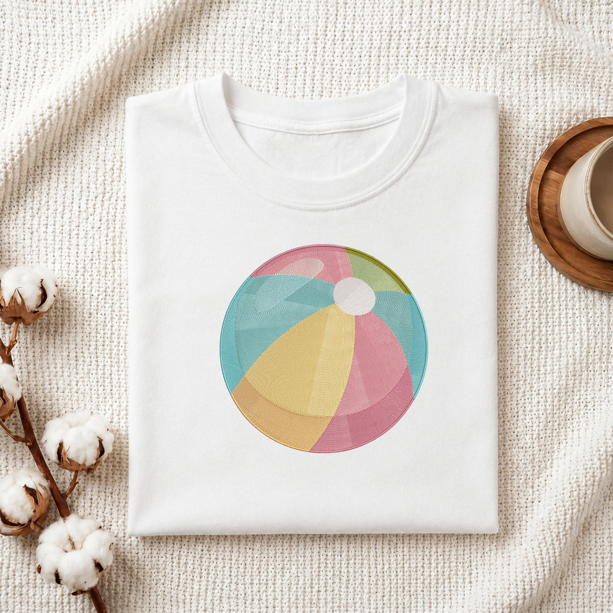 A folded white crewneck t-shirt with a colorful beach ball graphic printed on the chest, styled on a textured cream knit blanket with cotton bolls and a wooden cup in the background.