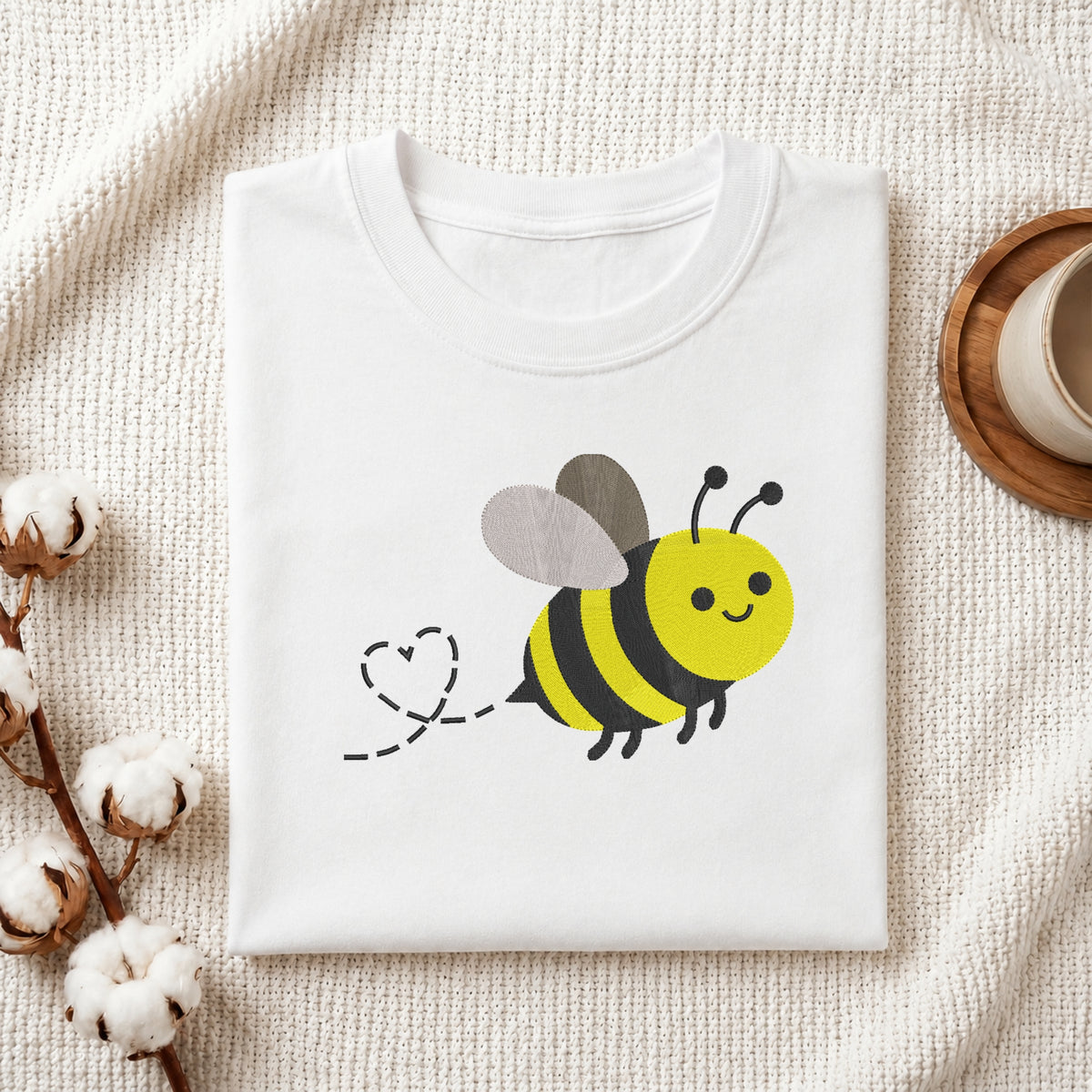 A folded white crewneck t-shirt featuring a cute cartoon bumblebee graphic with a dashed heart trail, styled on a textured cream knit blanket with cotton bolls and a wooden cup in the background.