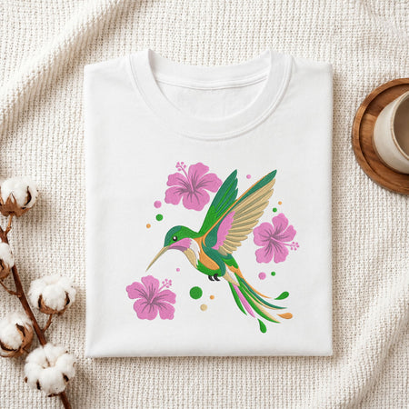A folded white crewneck t-shirt with a colorful hummingbird and pink hibiscus flower graphic, styled on a textured cream fabric with cotton bolls and a cup in the background.