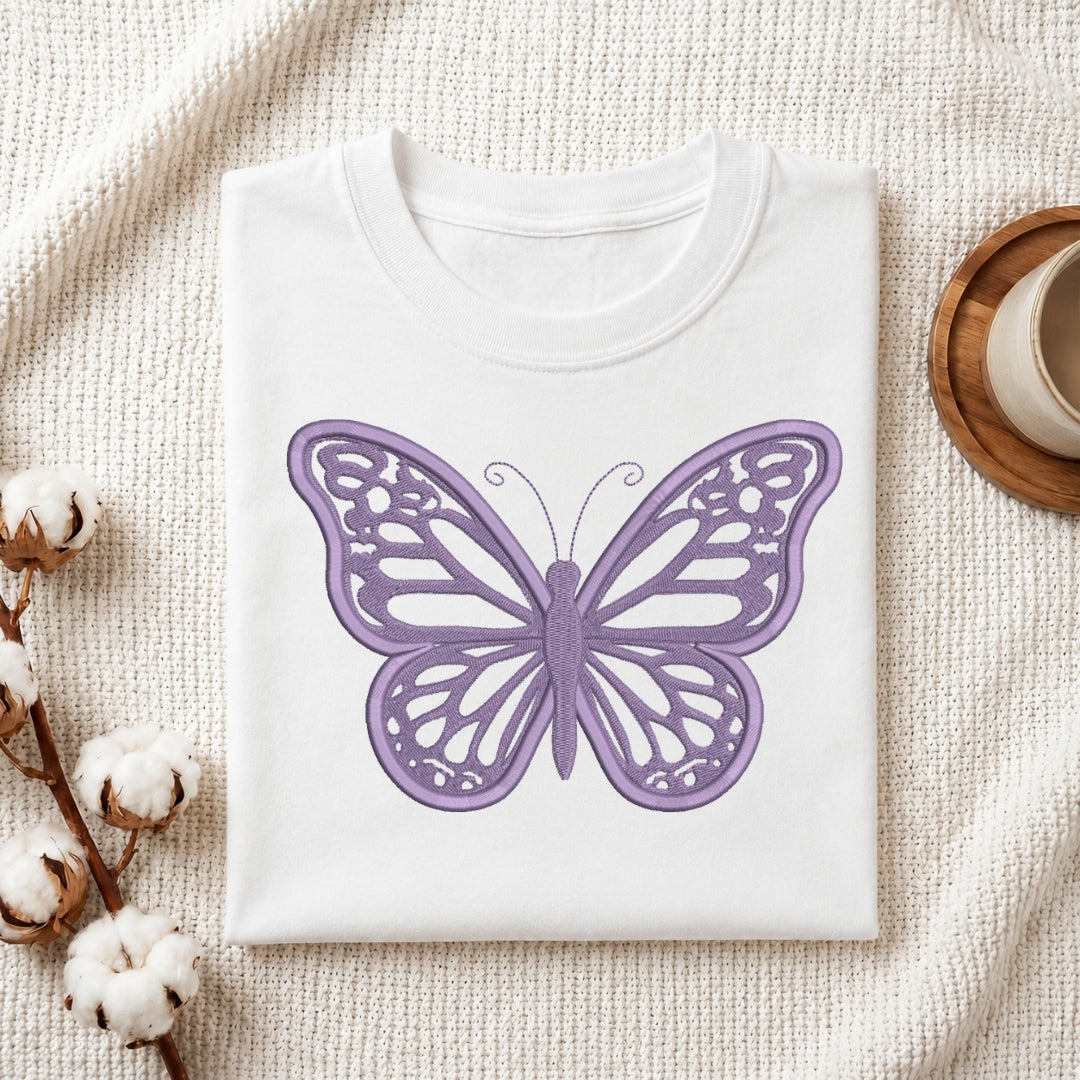 A folded white crewneck t-shirt with a purple embroidered butterfly design, styled on a textured cream fabric background with cotton bolls and a wooden cup.