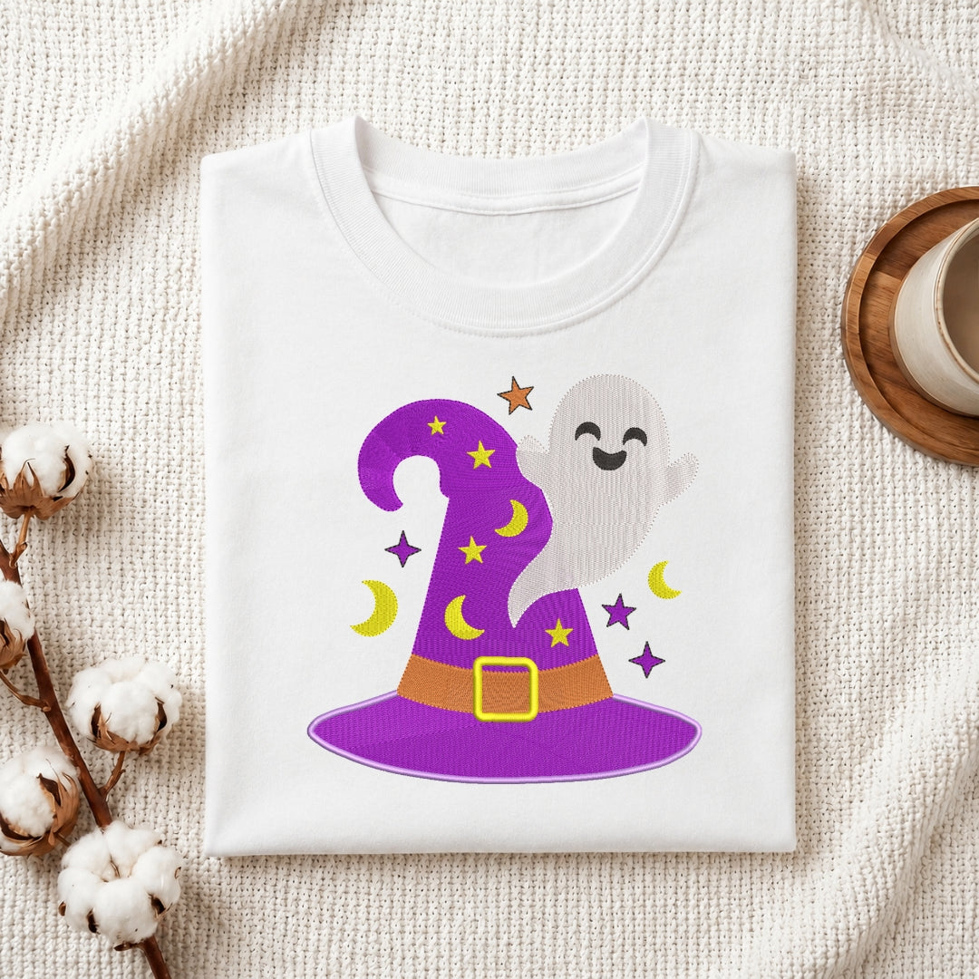 A folded white T-shirt or sweatshirt laid flat on a textured cream fabric, featuring a colorful Halloween graphic of a purple witch hat, a smiling ghost, yellow crescent moons, and stars. Decorative cotton stems and a cup on a coaster are partially visible around the shirt.