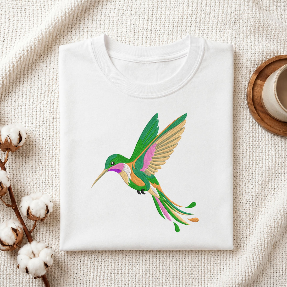 A folded white crewneck t-shirt featuring a colorful graphic of a hummingbird in green, pink, and gold, styled on a textured cream fabric with cotton bolls and a wooden cup.