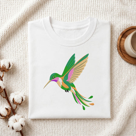 A folded white crewneck t-shirt featuring a colorful graphic of a hummingbird in green, pink, and gold, styled on a textured cream fabric with cotton bolls and a wooden cup.