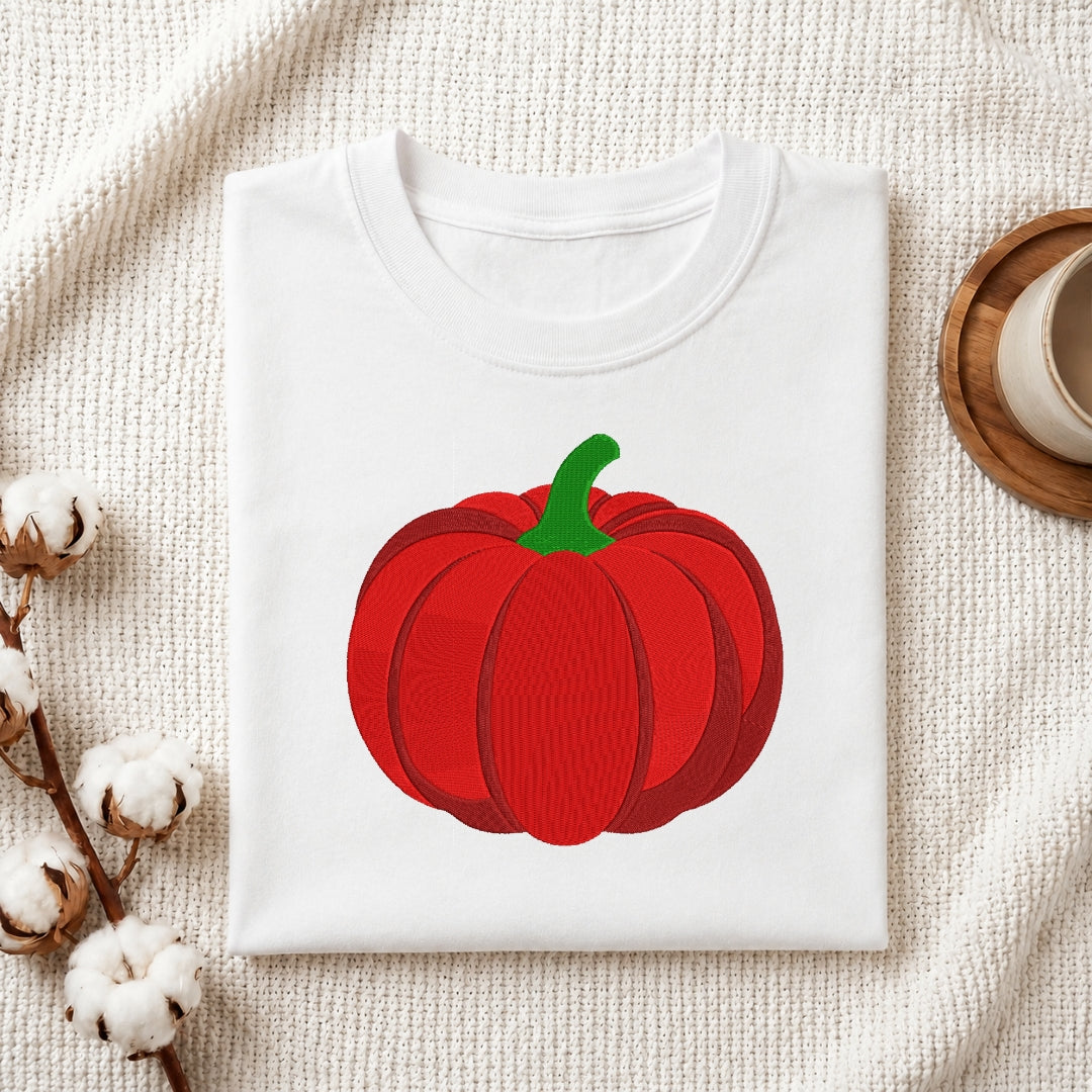 A folded white crewneck t-shirt featuring a graphic of a red pumpkin with a green stem, styled on a textured cream fabric with cotton bolls and a wooden cup in the background.