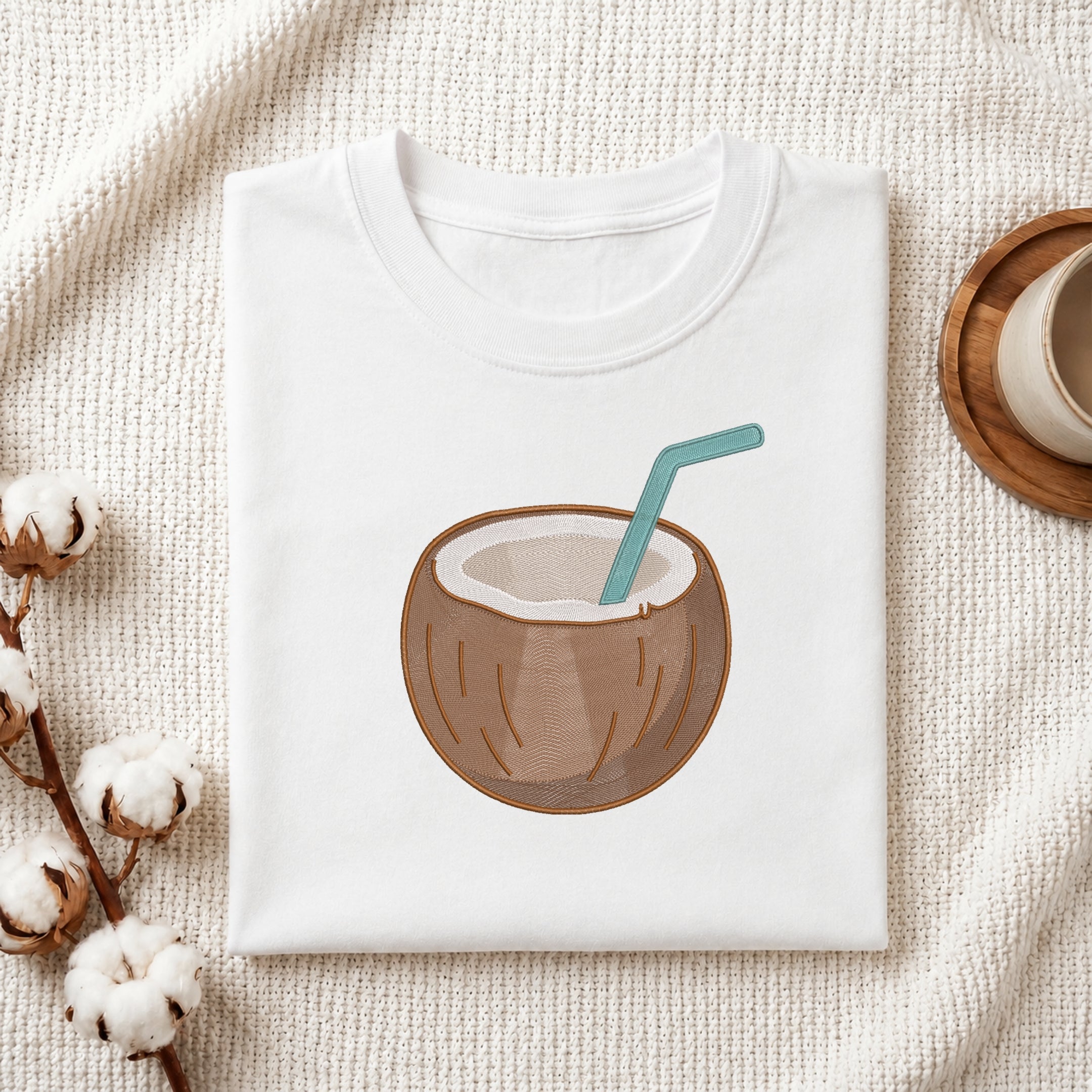 A folded white t-shirt with a graphic of a coconut drink in a brown coconut shell with a teal straw, styled on a textured cream fabric with cotton bolls and a wooden cup.