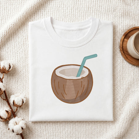 A folded white t-shirt with a graphic of a coconut drink in a brown coconut shell with a teal straw, styled on a textured cream fabric with cotton bolls and a wooden cup.