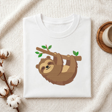 A folded white crewneck t-shirt featuring a cartoon sloth hanging from a branch with green leaves, styled on a textured cream knit blanket with cotton bolls and a cup on a wooden tray.