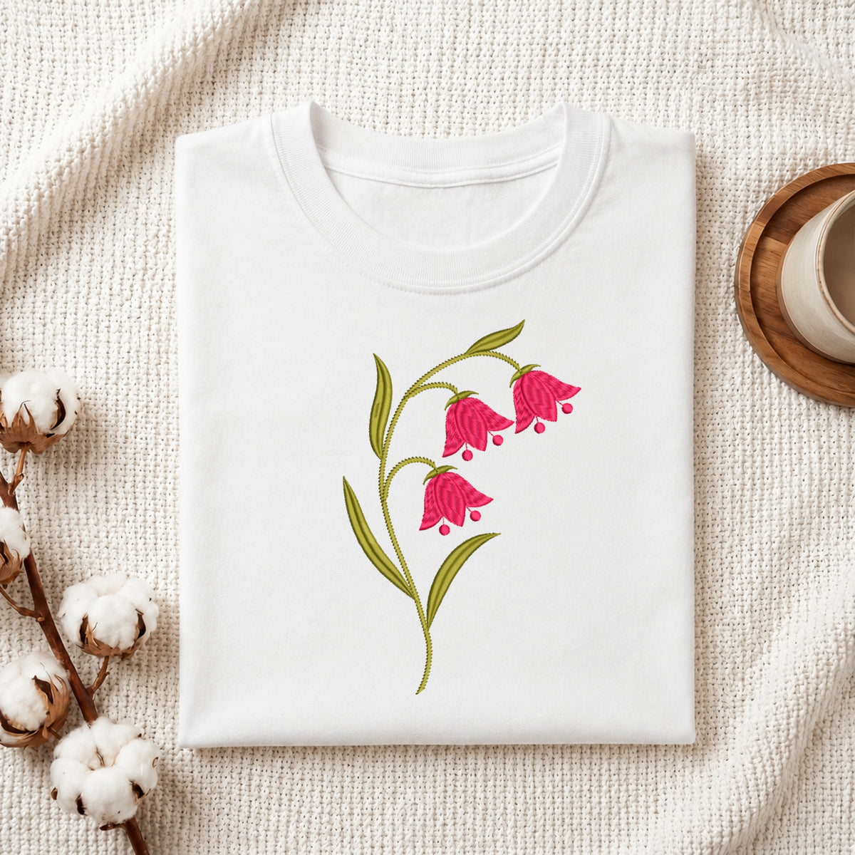 A folded white t-shirt with a pink bell-shaped flower and green leaves graphic, displayed on a textured cream fabric with cotton bolls and a wooden cup in the background.