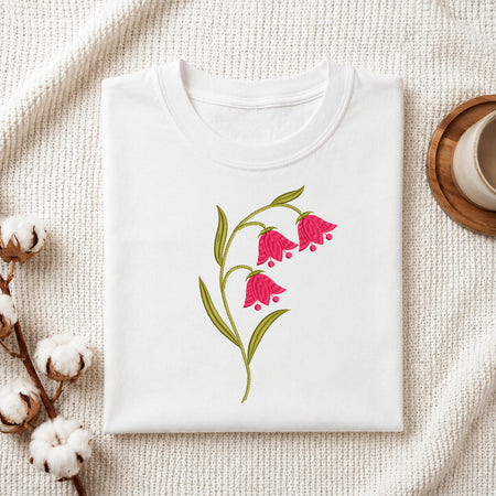 A folded white t-shirt with a pink bell-shaped flower and green leaves graphic, displayed on a textured cream fabric with cotton bolls and a wooden cup in the background.