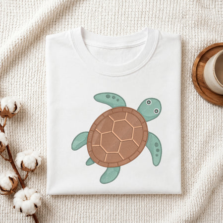 A folded white crewneck t-shirt featuring a cartoon sea turtle graphic on the front, styled on a textured cream fabric background with cotton bolls and a wooden cup.
