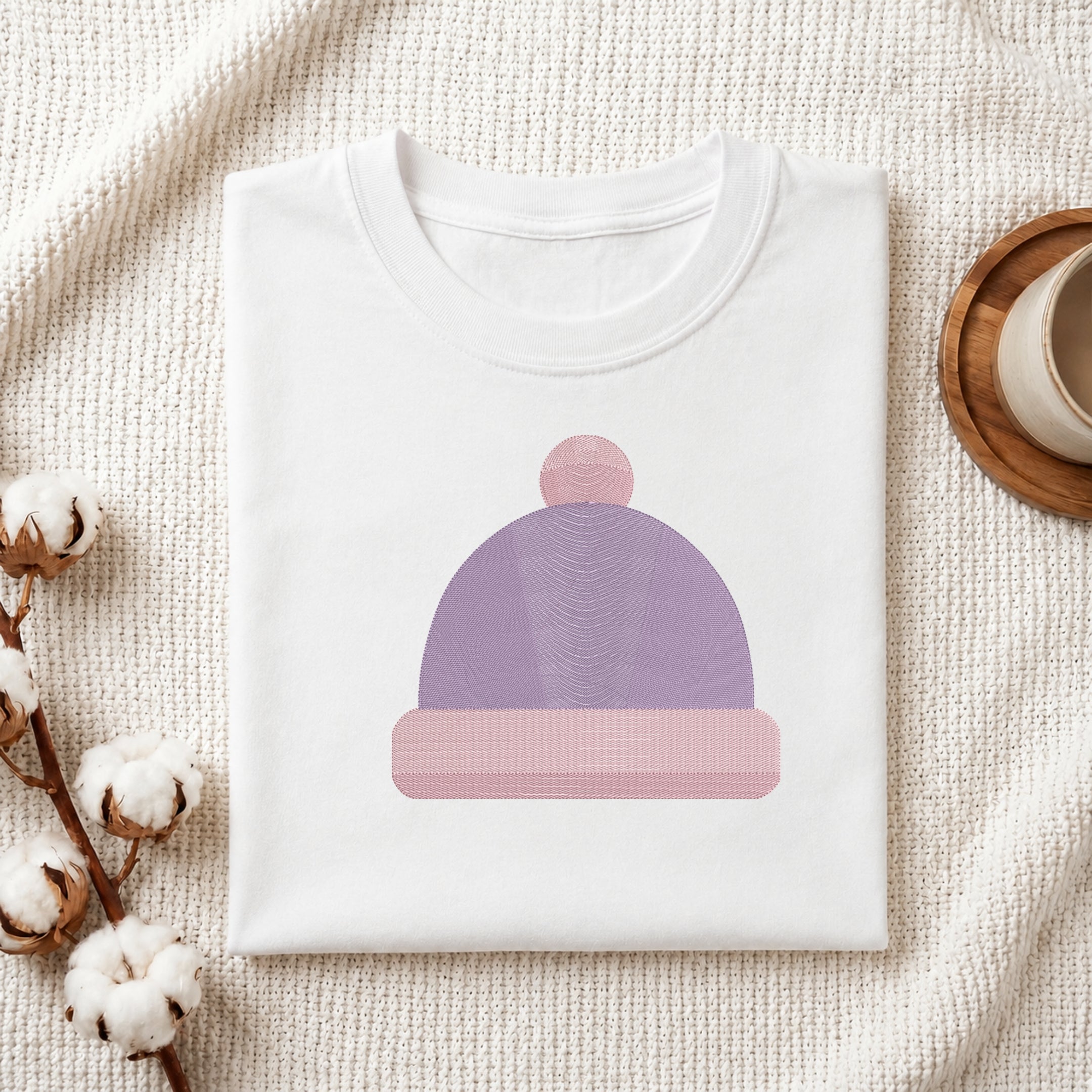 A folded white crewneck t-shirt featuring a stylized graphic of a lavender and pink beanie hat, styled on a textured cream fabric with cotton bolls and a wooden cup.
