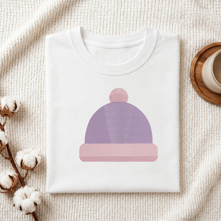 A folded white crewneck t-shirt featuring a stylized graphic of a lavender and pink beanie hat, styled on a textured cream fabric with cotton bolls and a wooden cup.