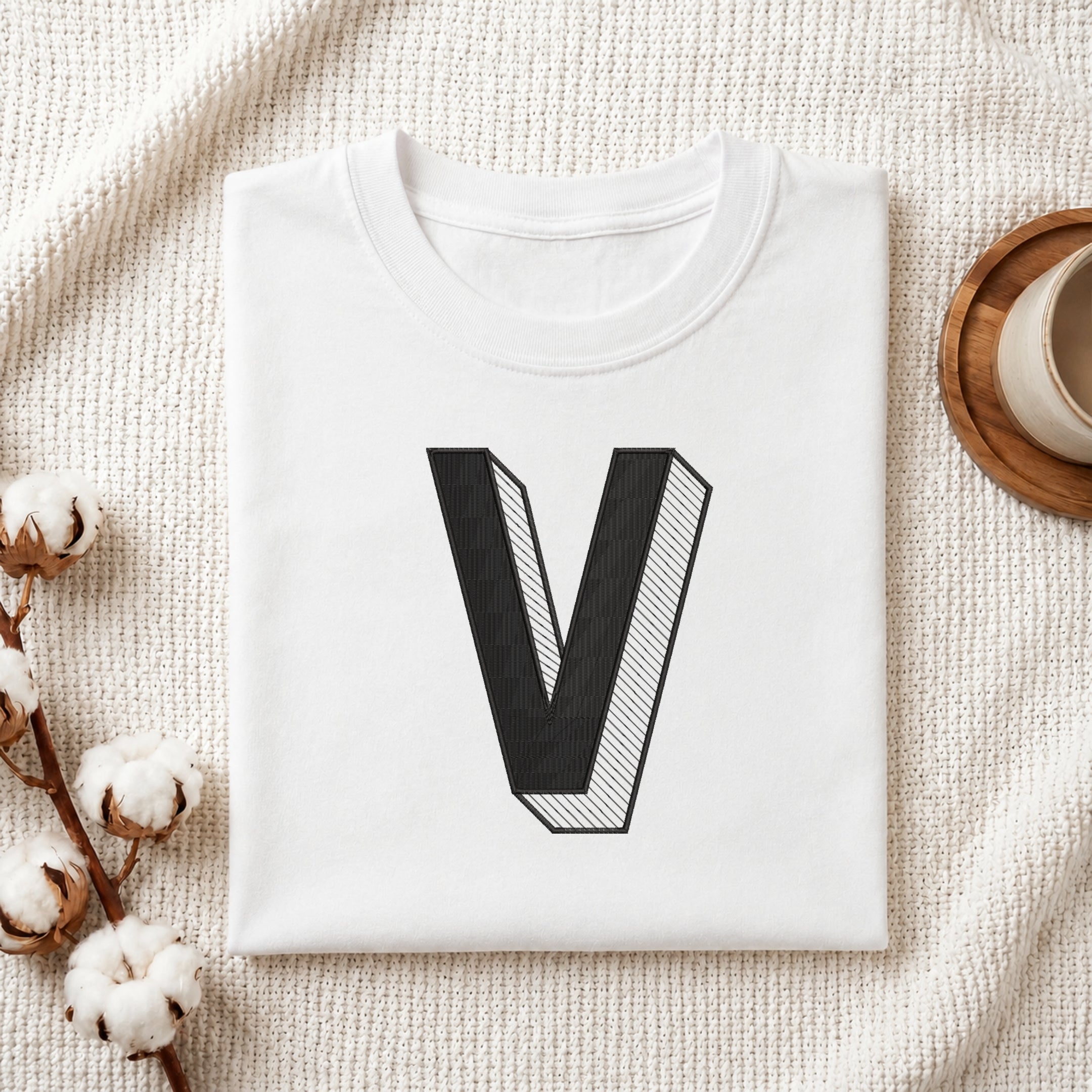 A folded white t-shirt with a large black 3D-style letter 'V' printed on the front, styled in a flat lay on a textured cream fabric with cotton bolls and a cup in the background.