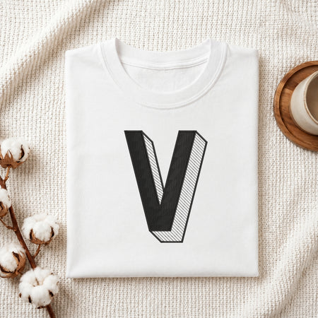 A folded white t-shirt with a large black 3D-style letter 'V' printed on the front, styled in a flat lay on a textured cream fabric with cotton bolls and a cup in the background.
