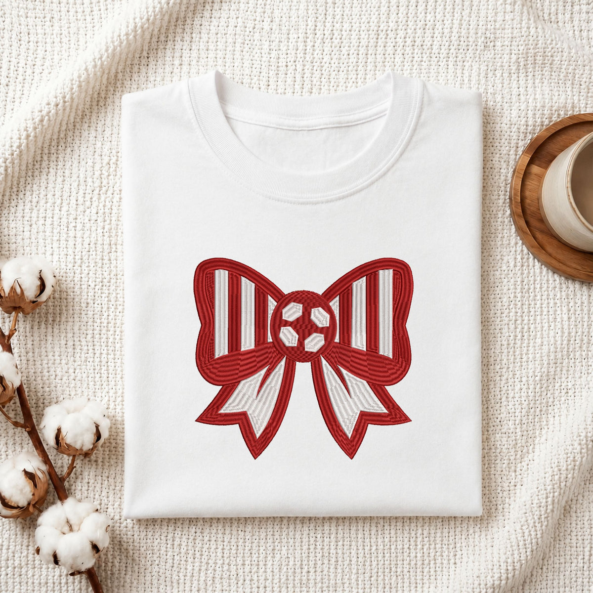 A folded white crewneck t-shirt with a red and white striped bow graphic on the front, styled on a textured cream blanket with cotton bolls and a wooden cup in the background.