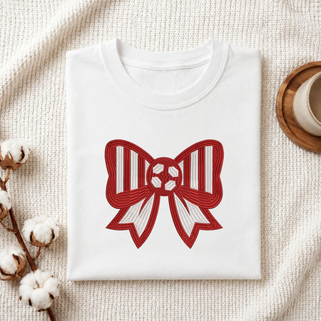 A folded white crewneck t-shirt with a red and white striped bow graphic on the front, styled on a textured cream blanket with cotton bolls and a wooden cup in the background.