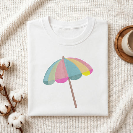 A folded white crewneck t-shirt with a pastel rainbow beach umbrella graphic, styled on a textured cream knit blanket with cotton bolls and a wooden cup in the background.