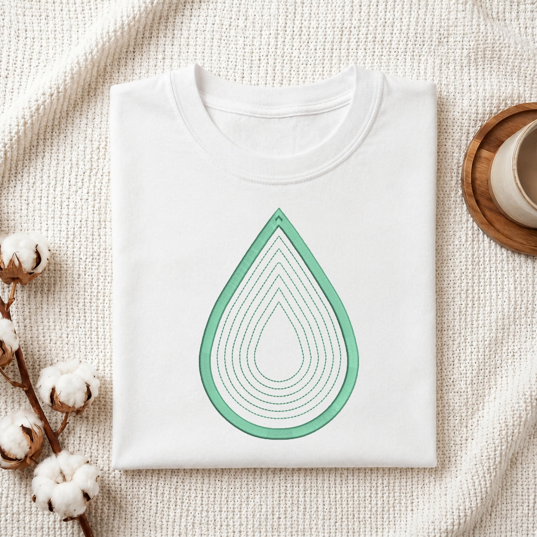 A folded white crewneck t-shirt with a mint green layered teardrop graphic on the front, styled on a textured cream knit blanket with cotton bolls and a wooden cup in the background.