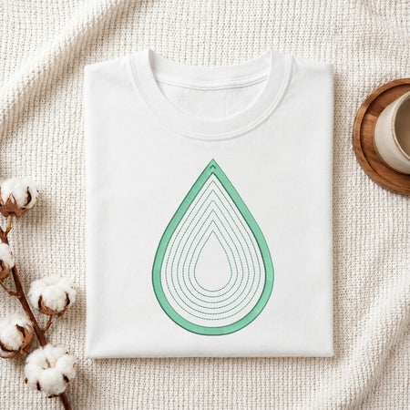 A folded white crewneck t-shirt with a mint green layered teardrop graphic on the front, styled on a textured cream knit blanket with cotton bolls and a wooden cup in the background.