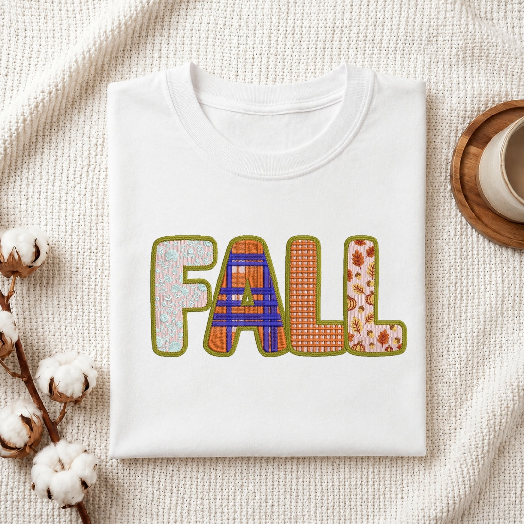 A folded white crewneck t-shirt with a colorful 'FALL' graphic printed on the front, styled on a textured cream knit blanket with cotton stems and a wooden cup in the background.