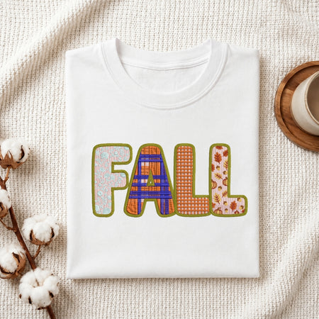 A folded white crewneck t-shirt with a colorful 'FALL' graphic printed on the front, styled on a textured cream knit blanket with cotton stems and a wooden cup in the background.