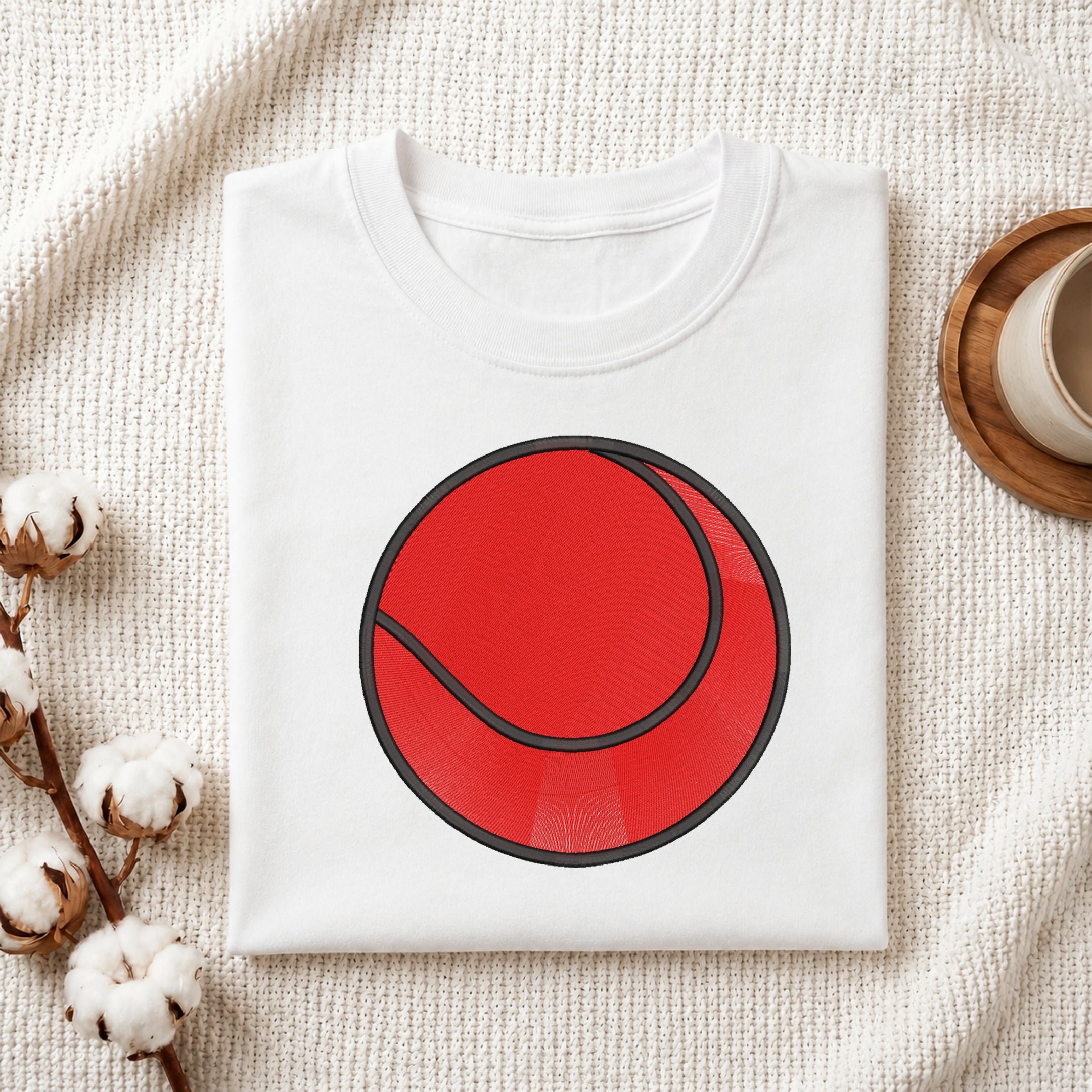 A folded white crewneck t-shirt featuring a large red tennis ball graphic on the front, styled on a textured cream fabric with cotton bolls and a wooden cup in the background.