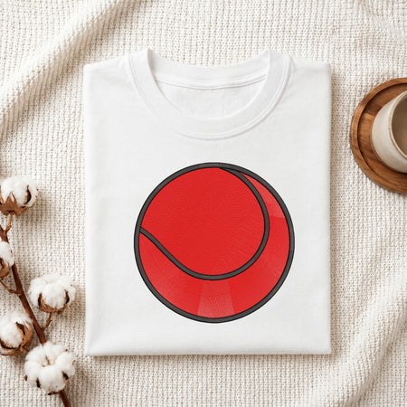 A folded white crewneck t-shirt featuring a large red tennis ball graphic on the front, styled on a textured cream fabric with cotton bolls and a wooden cup in the background.