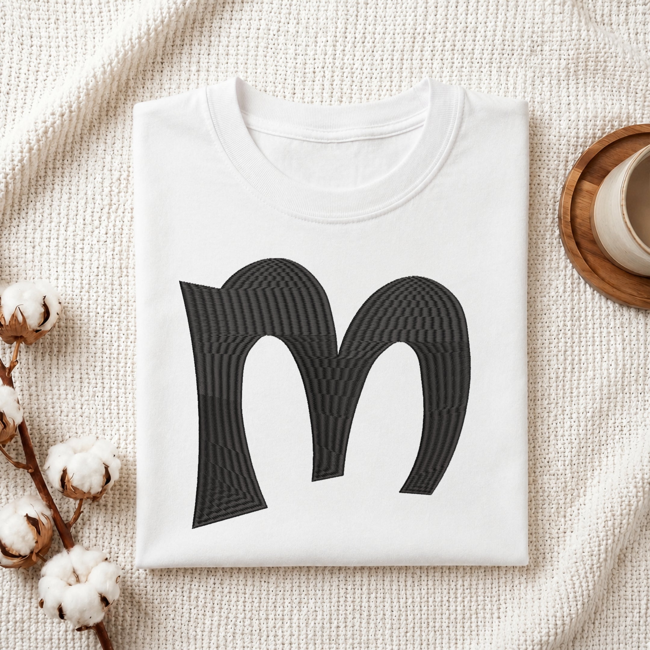 A folded white crewneck t-shirt with a large black stylized letter 'm' printed on the front, styled on a textured cream knit blanket with cotton bolls and a wooden cup in the background.