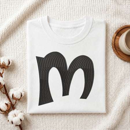 A folded white crewneck t-shirt with a large black stylized letter 'm' printed on the front, styled on a textured cream knit blanket with cotton bolls and a wooden cup in the background.