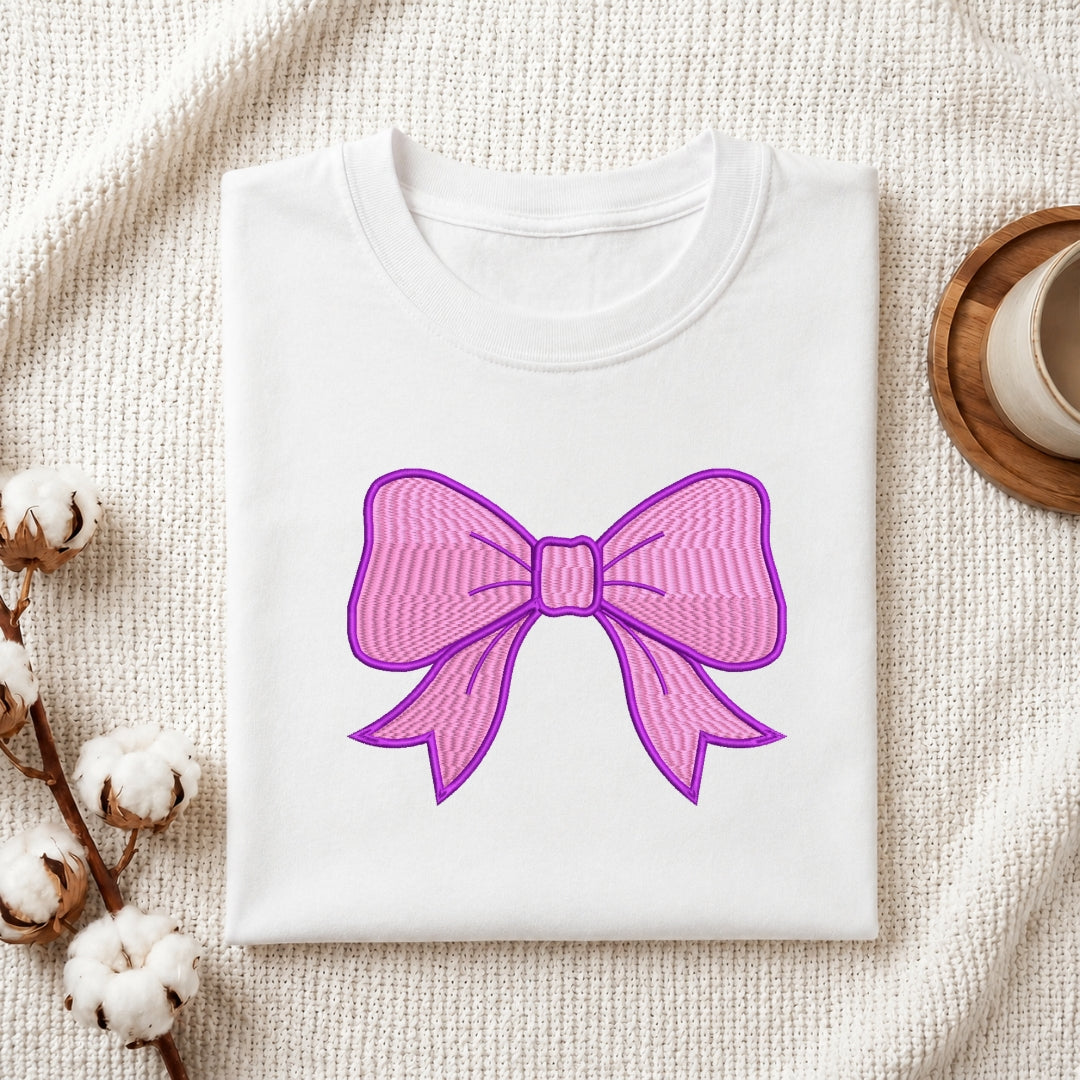 A folded white crewneck t-shirt with a pink and purple striped bow graphic on the front, styled on a textured cream knit blanket with cotton bolls and a wooden cup in the background.