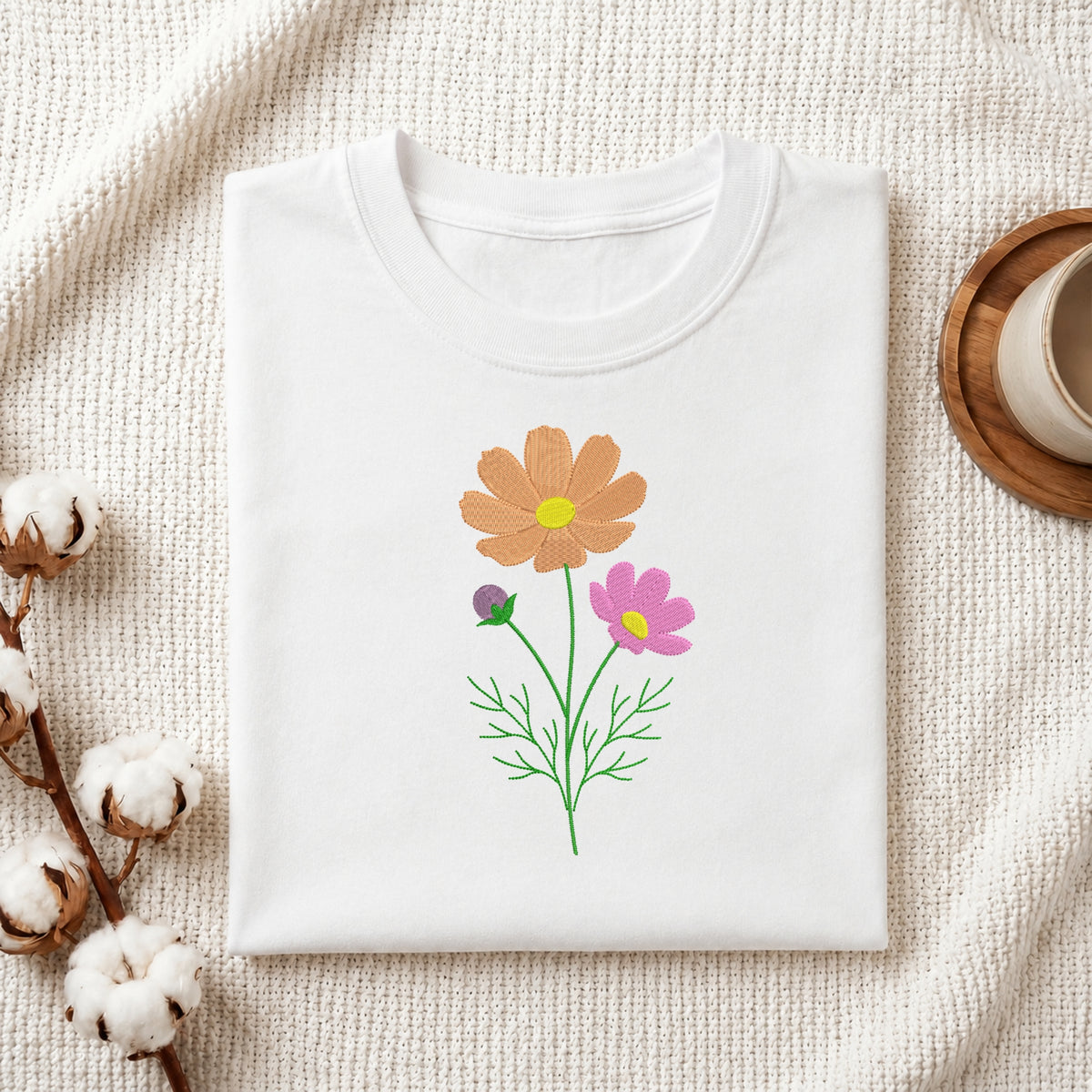 A folded white crewneck t-shirt with a colorful floral graphic of two flowers (one orange, one pink) and green stems, styled on a textured cream fabric with cotton bolls and a wooden cup.