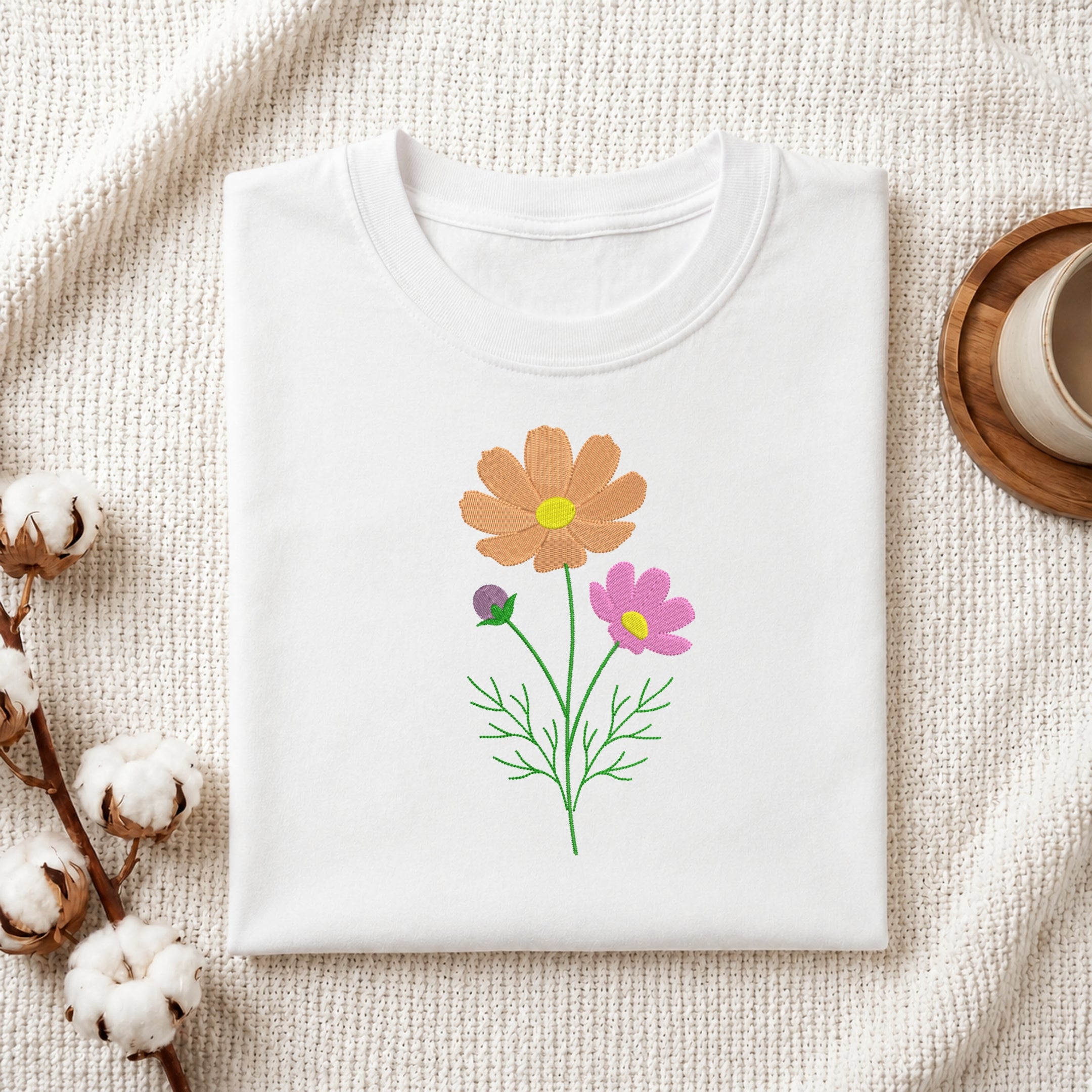 A folded white crewneck t-shirt with a colorful floral graphic of two flowers (one orange, one pink) and green stems, styled on a textured cream fabric with cotton bolls and a wooden cup.