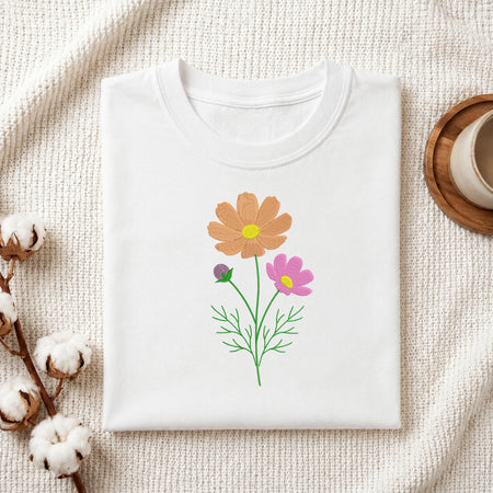 A folded white crewneck t-shirt with a colorful floral graphic of two flowers (one orange, one pink) and green stems, styled on a textured cream fabric with cotton bolls and a wooden cup.