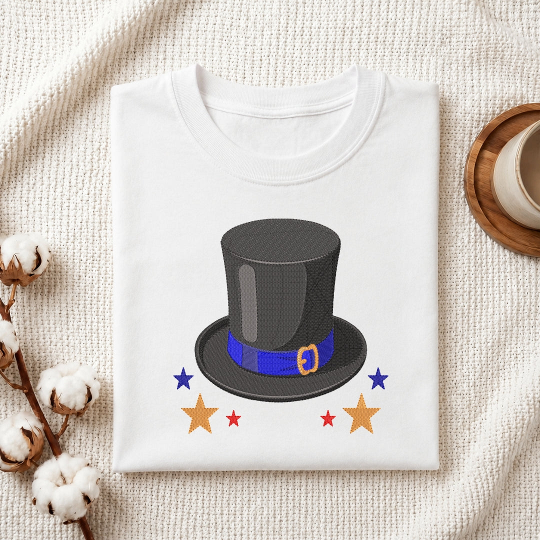 A folded white crewneck t-shirt with a graphic of a black top hat with a blue band and gold buckle, surrounded by stars, presented on a textured cream fabric background with decorative cotton bolls and a coffee cup.