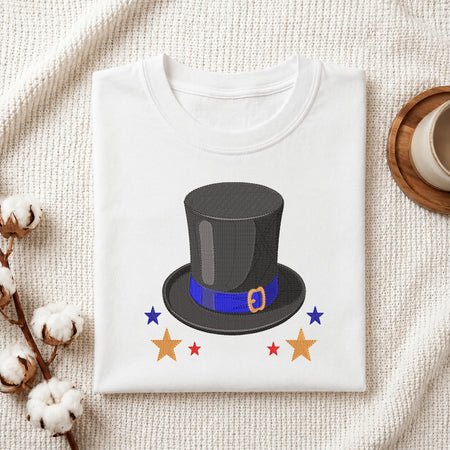 A folded white crewneck t-shirt with a graphic of a black top hat with a blue band and gold buckle, surrounded by stars, presented on a textured cream fabric background with decorative cotton bolls and a coffee cup.
