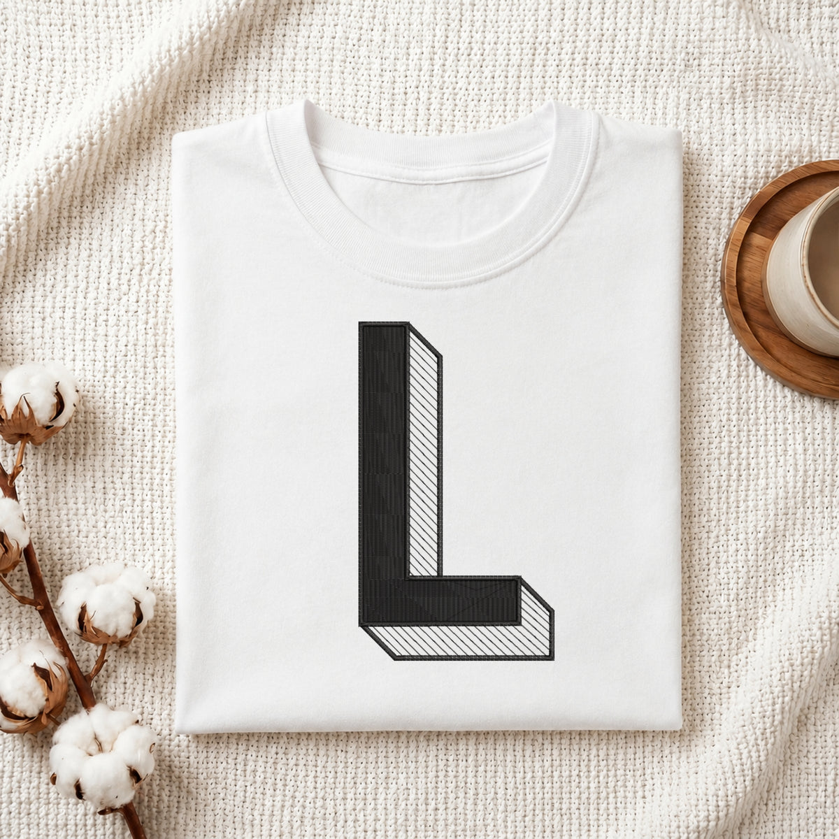 A folded white T-shirt with a large black block letter 'L' printed on the front, styled on a textured cream fabric with cotton stems and a cup in the background.